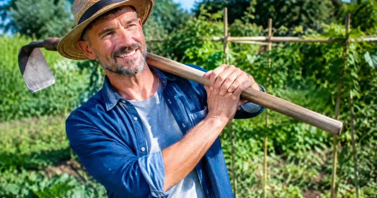 Jardinier tenant faux courbée sur son epaule dans potager verdoyant